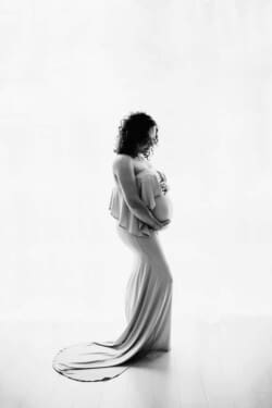 A maternity image of a mother in a flowing gown stands in front of a large window holding her pregnant belly