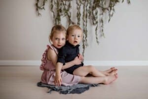A little girl holds her baby brother in her lap as they sit on a white timber floor for his birthday session