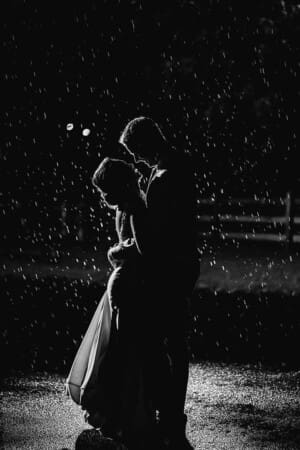 A groom hugs hugs bride at night with a bright light behind them as rain falls