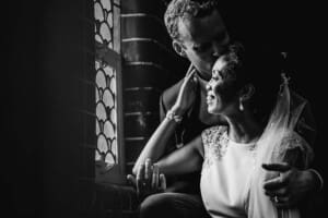 A groom kisses his bride on the cheek as they sit next to a church window after the ceremony
