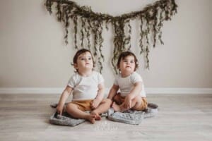Twin babies sit on a grey blanket in a bright studio with a green leafy backdrop