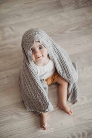 A smiling baby boy sits on a white wood floor with a grey blanket draped over him