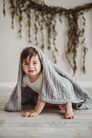 A smiling baby boy crawls across a white wood floor with a grey blanket draped over him