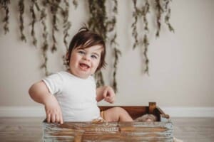 A smiley baby boy in a white romper sits in a wooden crate with a green leafy backdrop