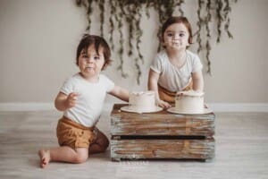 Twin boys sit beside a wooden crate and smash their first birthday cakes