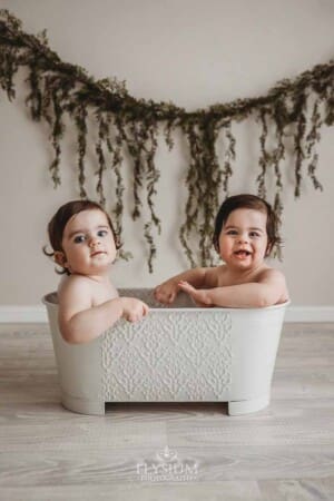 Twin boys sit in a white bathtub in a bright studio with hanging leaves in the background