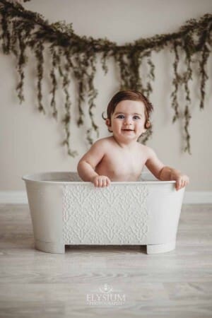 A baby boy smiles while sitting in a white vintage tub with a leafy backdrop