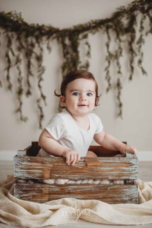 A smiley baby wearing a white romper sits in a wood crate with green leaves in the background
