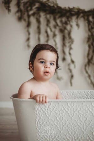 A baby boy sits in a white vintage bathtub and cleans off after his cake smash