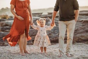 Parents hold their little girls' hands as she stands between them on a rocky beach