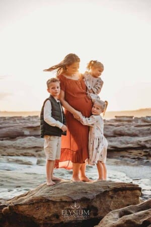 A pregnant woman hugs her three children as they stand on a beach rock