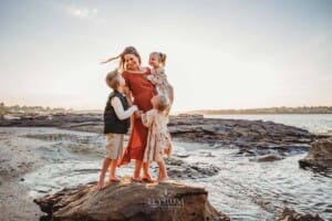 A pregnant woman hugs her three children as they stand on a beach rock