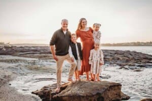 A family stand cuddling on a large beach rock at sunset