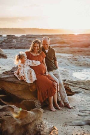 A little girl puts her hand on her pregnant mum's belly as they sit on a beach rock at sunset