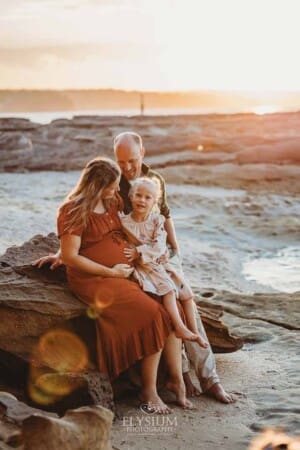 A girl puts her hand on her pregnant mum's belly as they sit on a beach rock at sunset