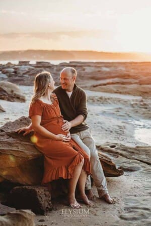 A husband holds his wife's pregnant belly as they sit on a beach rock at sunset
