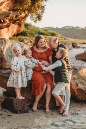 A pregnant woman sits on a beach rock with her family as her children place their hands on her belly