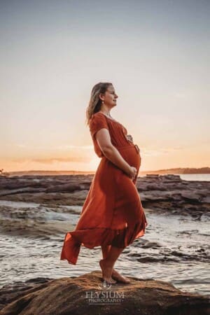 A pregnant woman stands on a rock at the beach with the colourful sunset behind her