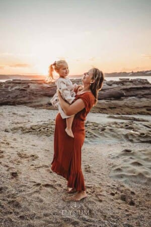 A pregnant mum holds her little girl as they stand on a sandy beach at sunset