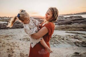 A pregnant mum holds her little girl as they stand on a sandy beach at sunset