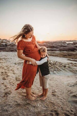 A boy cuddles his mum's pregnant belly as they stand on a sandy beach at sunset
