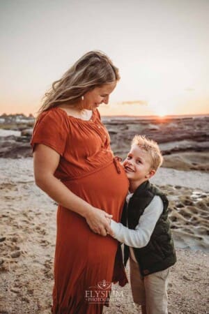 A boy cuddles his mum's pregnant belly as they stand on a sandy beach at sunset