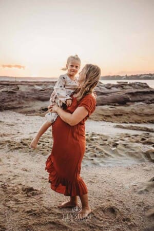 A pregnant mum holds her little girl as they stand on a sandy beach at sunset