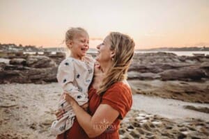A pregnant mum holds her little girl as they stand on a sandy beach at sunset