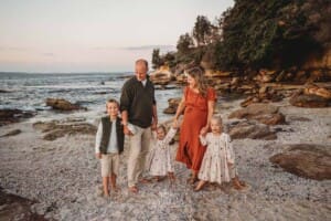 Parents stand on a sandy beach and hold hands with their three children at sunset