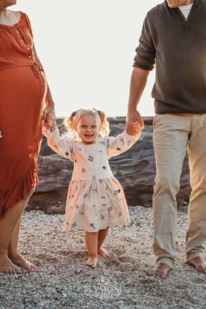 Parents hold their little girls hands as she stands between them on a beach