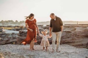 Parents hold their little girls hands as she stands between them on a sandy beach