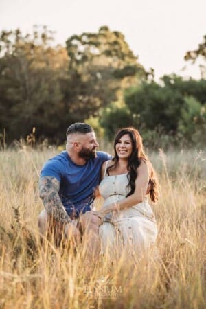 A man sits on a log with his pregnant wife amongst long grass at sunset