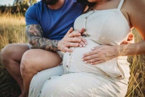 A couple sit holding the mum's pregnant belly as she wears a cream maternity dress