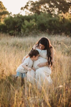 A little boy sits with his mum in a grassy field and kisses her pregnant belly