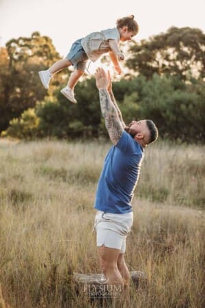 A dad lifts his son into the air as they stand in a grassy field at sunset