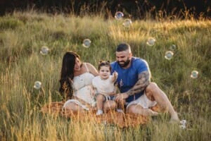 A little boy sits on a blanket with his dad and pregnant mum in a grass field with bubbles surrounding them