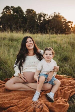 A little boy smiles sitting in his pregnant mum's lap surrounded by long grass
