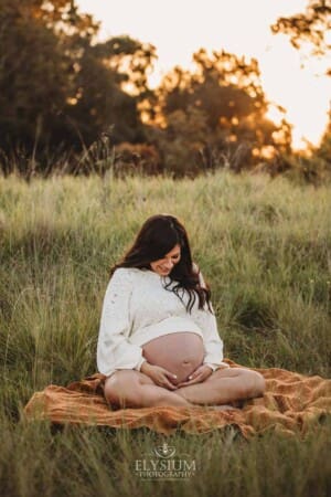 A pregnant woman wearing a white cropped jumper sits on an orange blanket in a grassy field at sunset