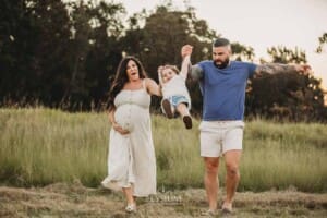 A pregnant woman and her husband walk through a grassy field and swing their little boy between them
