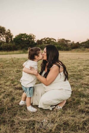 A pregnant mother crouches down to kiss her little boy as they stand in a field at sunset