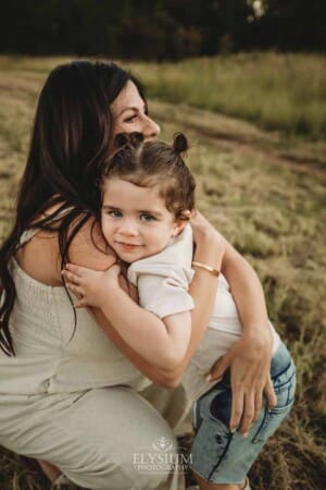 A little boy cuddles his pregnant mum as they stand in a grassy sundrenched field