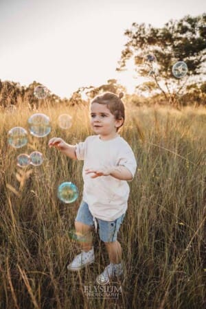 A little boy chases bubbles through long grass at sunset
