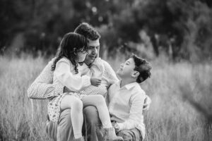 A father and children sit closely together in a grassy field at Ingleburn, captured during a relaxed sunset outdoor family session