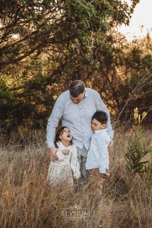 Children cuddled close to their dad as the sun dips behind the trees, a heartfelt outdoor family photography session in Ingleburn
