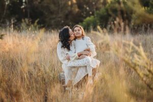 A mother kisses her child's cheek as they sit in long grass, a warm and natural family photography scene at golden hour