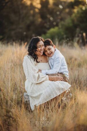 A boy sits in his mum's lap cuddling in long grass, an enjoyable and natural family photography moment at sunset.