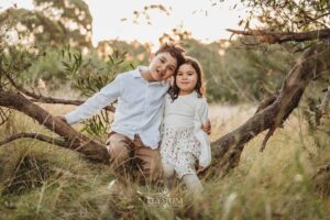 Children cuddled close sitting on a tree branch as the sun dips behind the trees, a heartfelt outdoor family photography moment