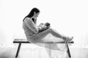 Mother sitting on a wood bench holding her newborn in gentle natural light inside the Ingleburn studio